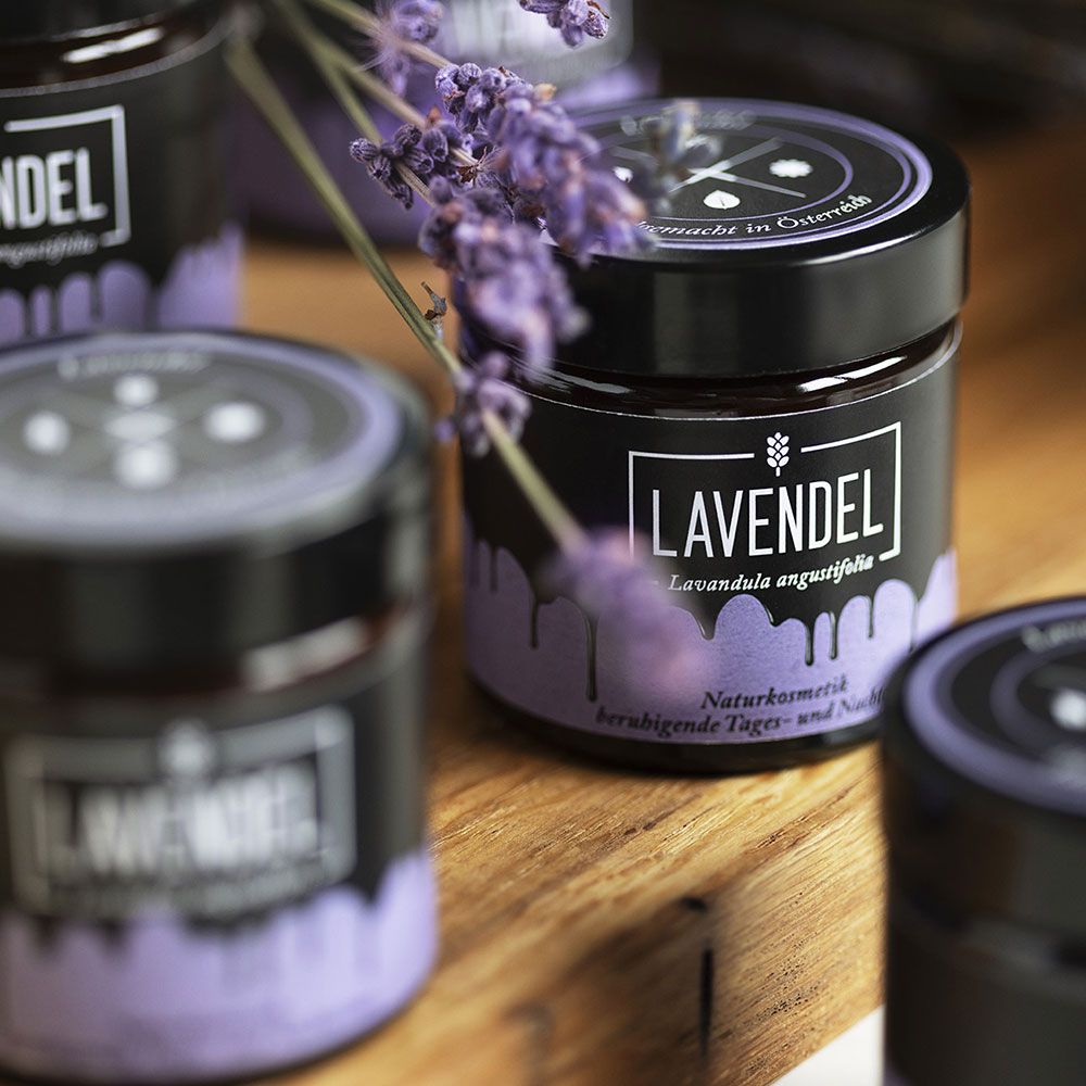 Mehrere Gläser mit schwarzem Etikett und Lavendel-Schriftzug. Lavendelzweige liegen auf Holz.