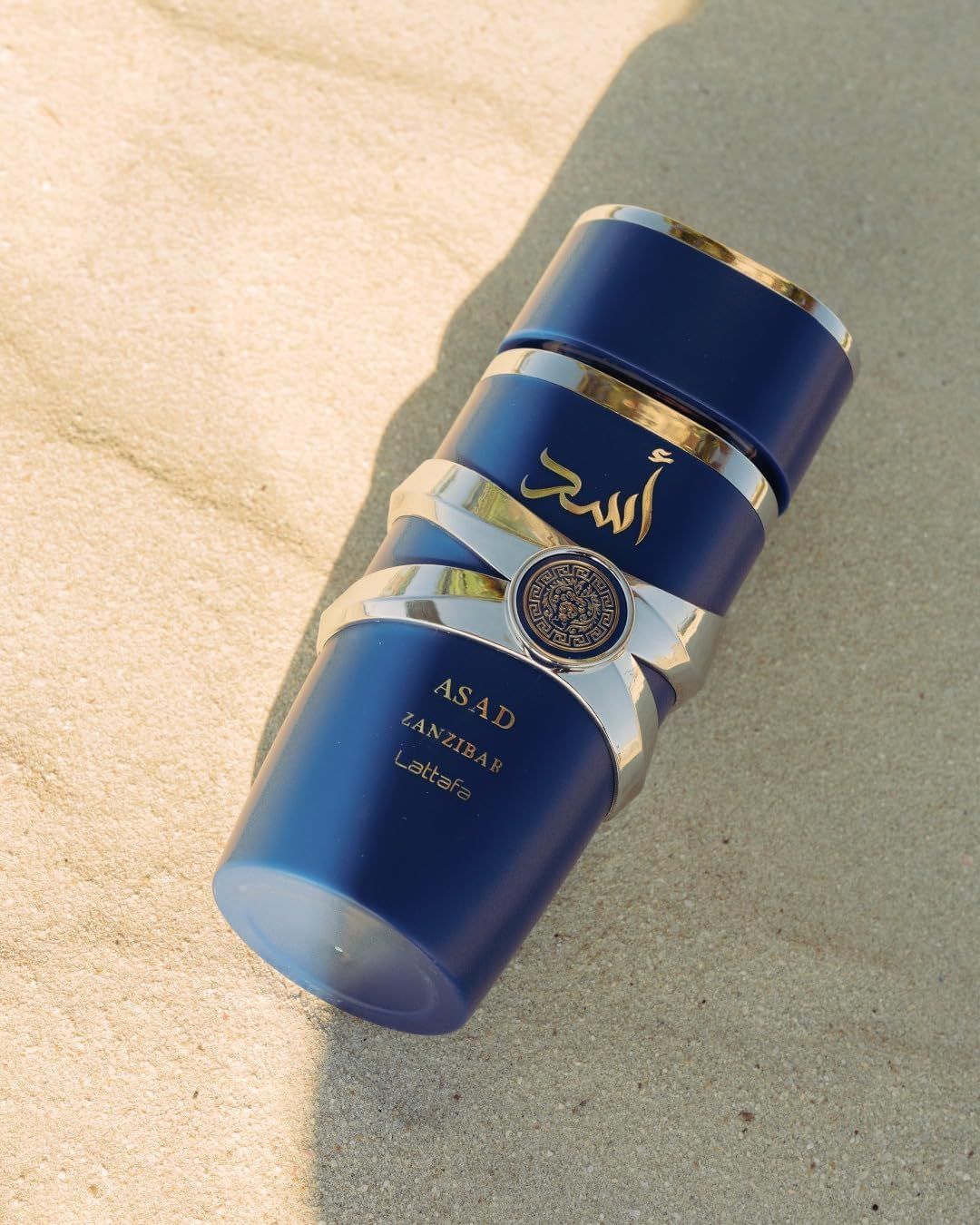 Blauer Flakon mit silbernen Bändern und Logo. Aufschrift: ASAD, ZANZIBAR, Lattafa. Liegt auf Sand. Goldfarbene Details.