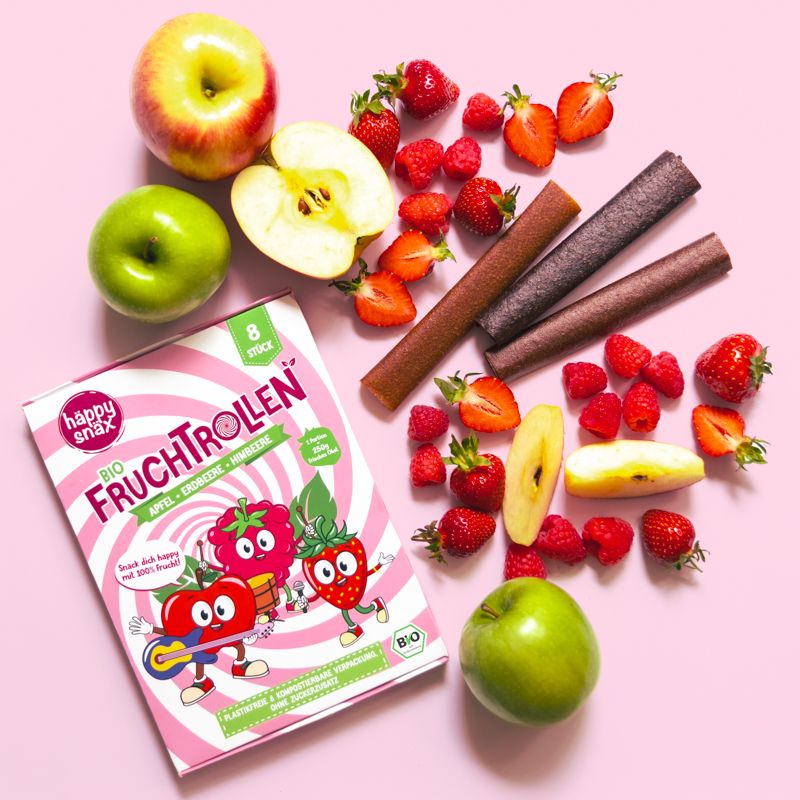 BIO Fruchtrollen und frische Früchte auf rosa Untergrund. Äpfel, Erdbeeren und Himbeeren sind sichtbar.