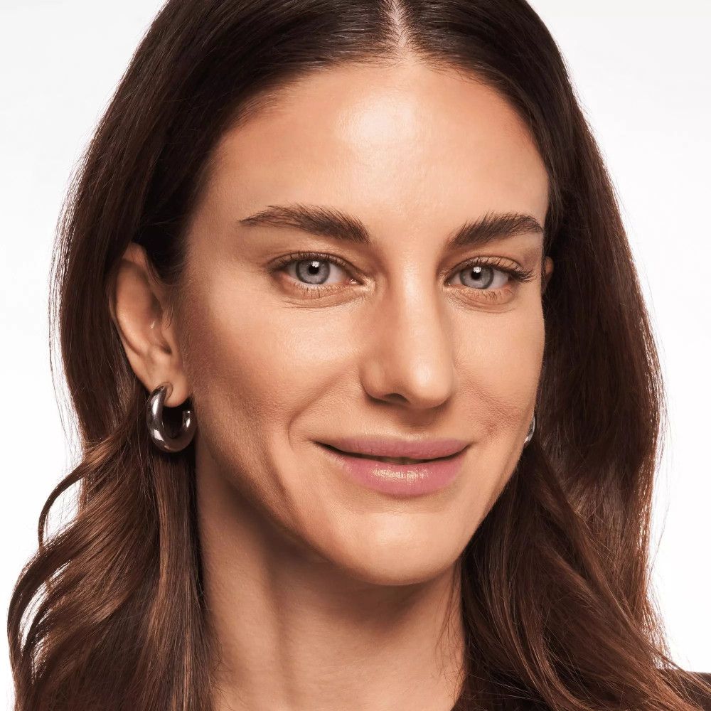 Porträt einer Frau mit braunen Haaren und silbernen Creolen. Sie lächelt.