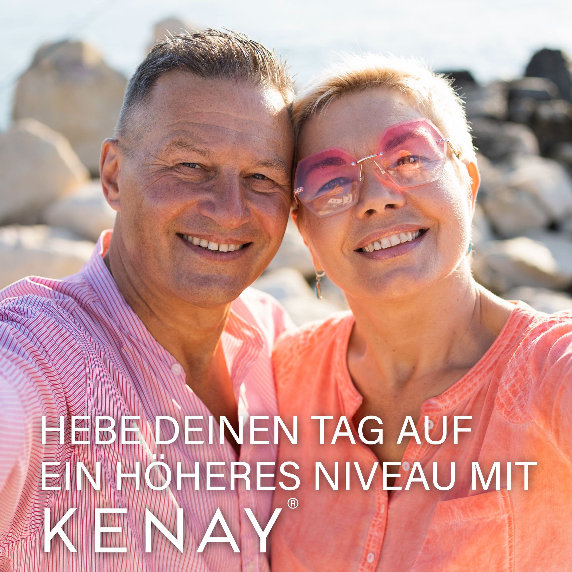 Zwei lächelnde Personen, Mann und Frau, im Freien. Text: HEBE DEINEN TAG AUF EIN HÖHERES NIVEAU MIT KENAY®. Sonnenbrille, rosa Hemd.
