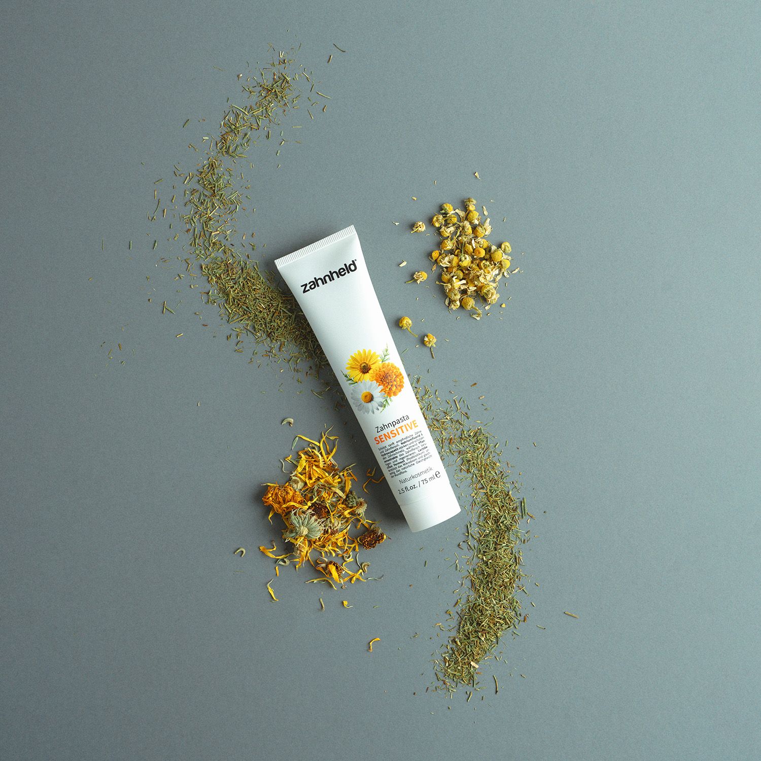 Zahnheld Zahnpasta Sensitive Tube. Kräuter und Blüten umgeben die Tube. Aufschrift: Zahnheld, Sensitive.