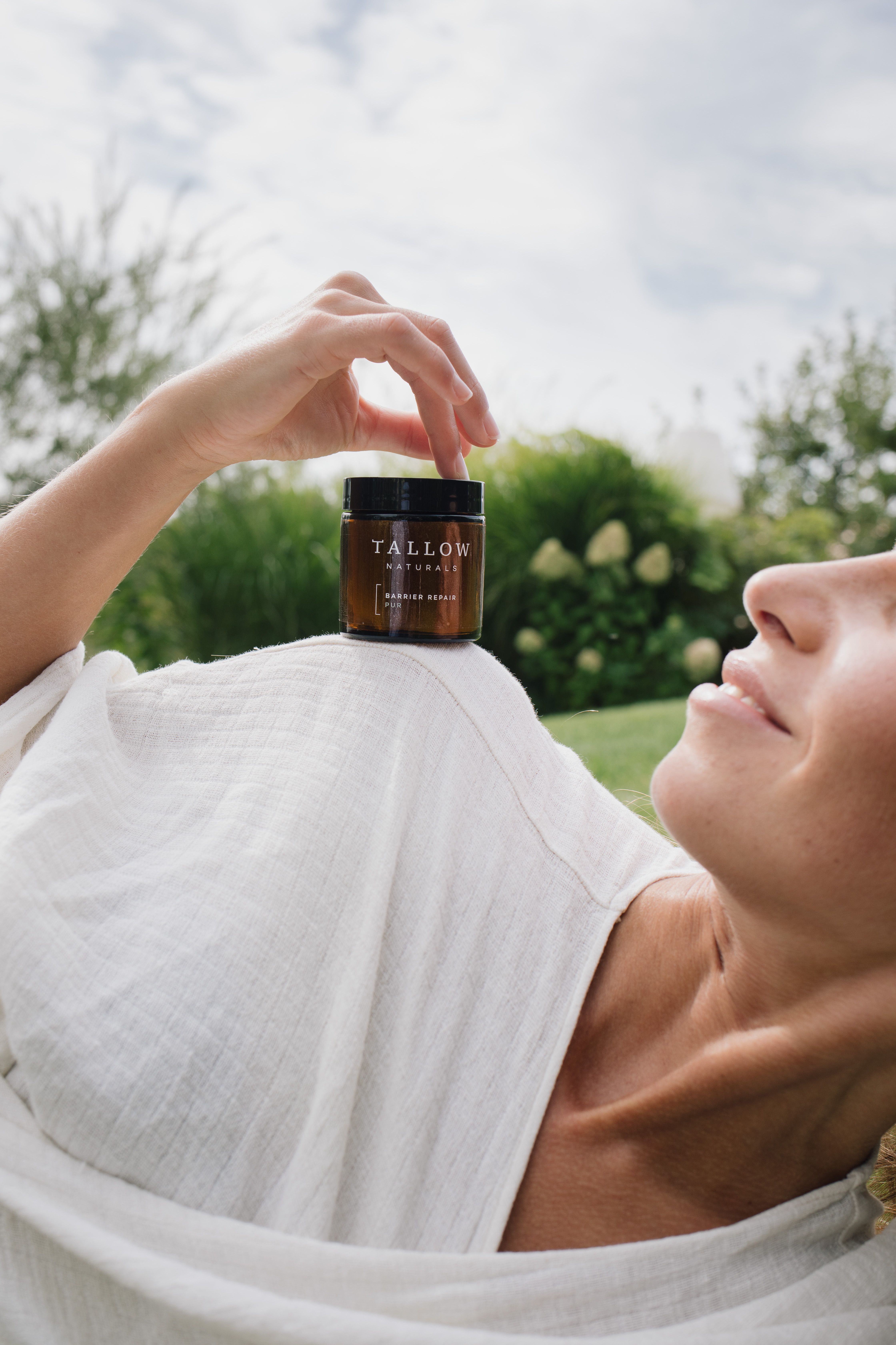 Frau liegt im Freien und hält eine braune Dose mit schwarzem Deckel. Aufschrift: Tallow Naturals, Barrier Repair Pur.
