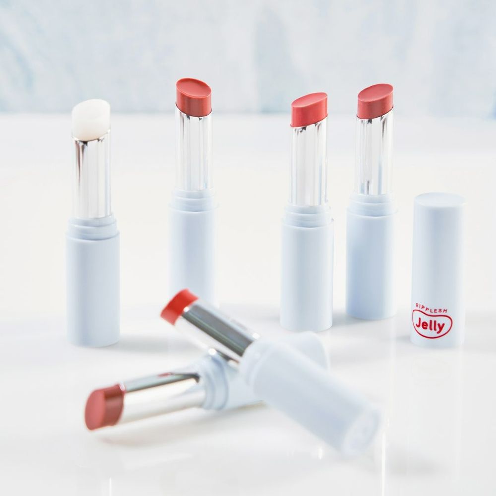 Mehrere Lippenbalsam-Stifte, einige geöffnet, in verschiedenen Farben. Weiße Behälter mit rotem Aufdruck "Ripplesh Jelly".