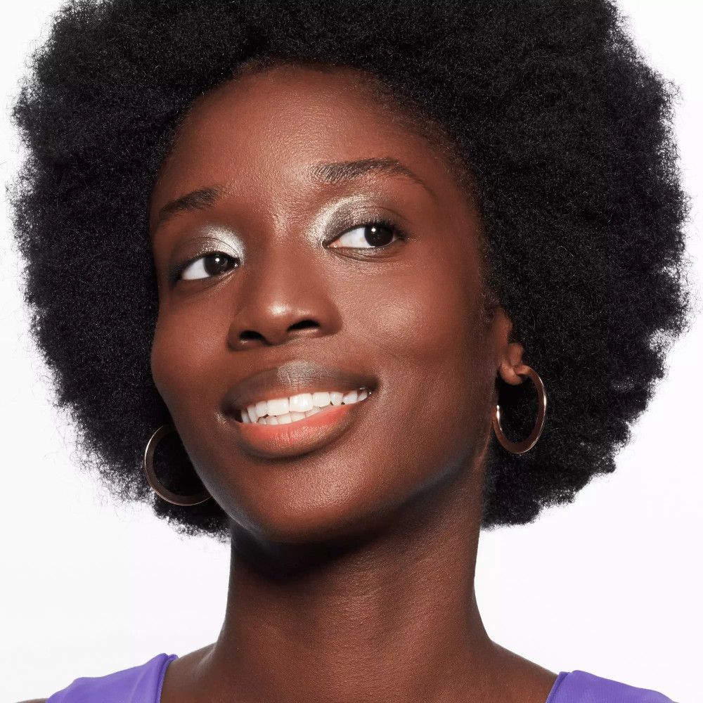 Porträt einer Frau mit dunkler Haut und silbernem Lidschatten. Sie lächelt und trägt goldene Creolen.