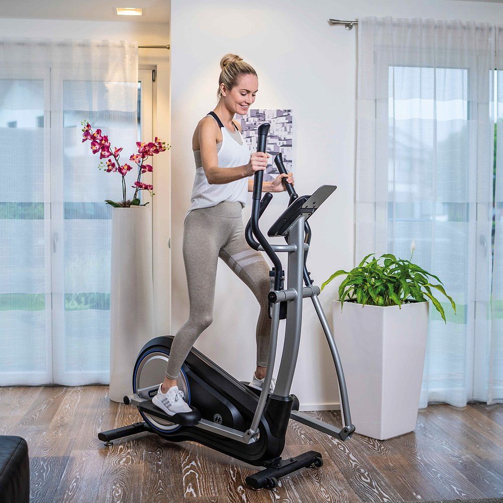 Frau trainiert auf dem Horizon Fitness Milos 3.0 Crosstrainer. In Wohnraum. Graue Sportkleidung. Pflanze im Hintergrund.