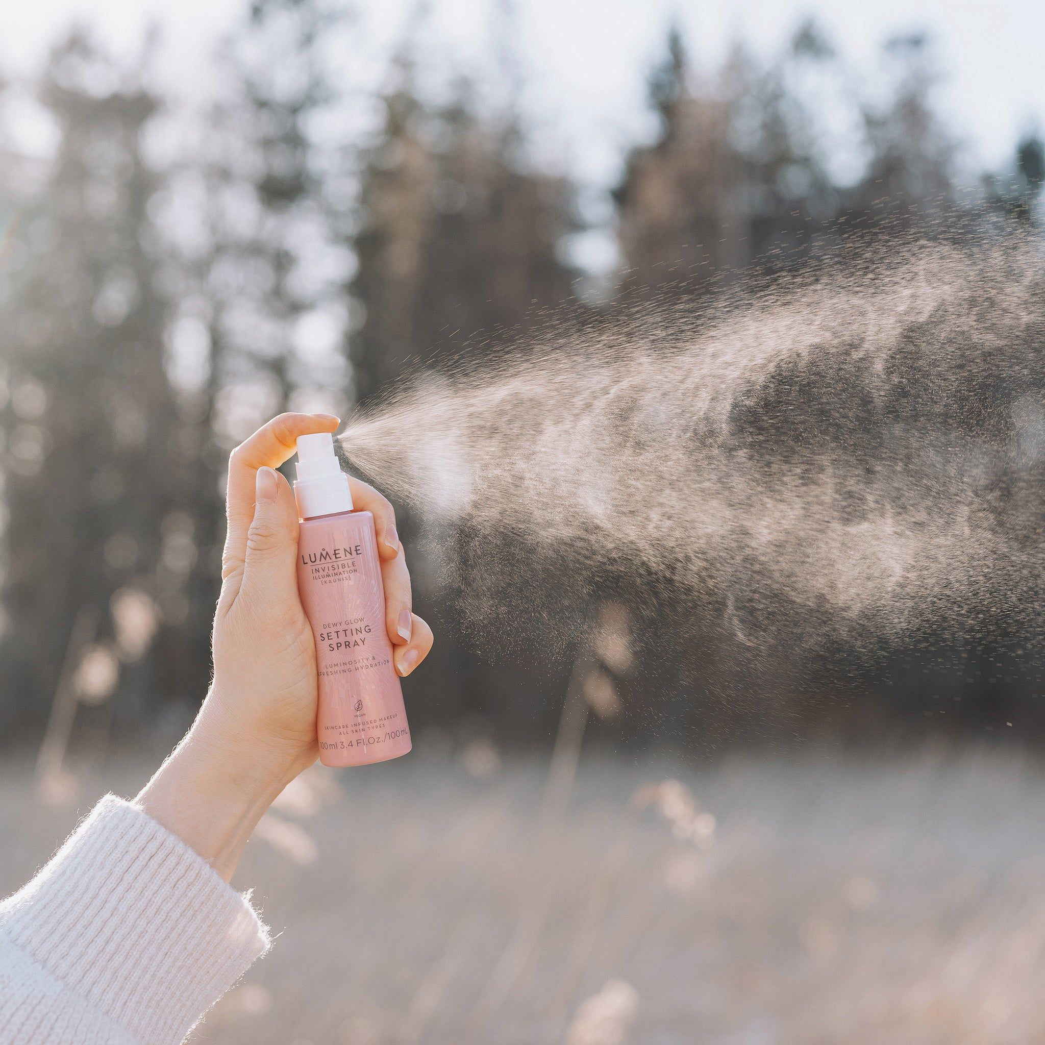 Hand hält rosa Sprühflasche. Sprühnebel. Aufschrift: Lumene INVISIBLE ILLUMINATION Dewy Glow Setting Spray. Hintergrund: Natur, Bäume.