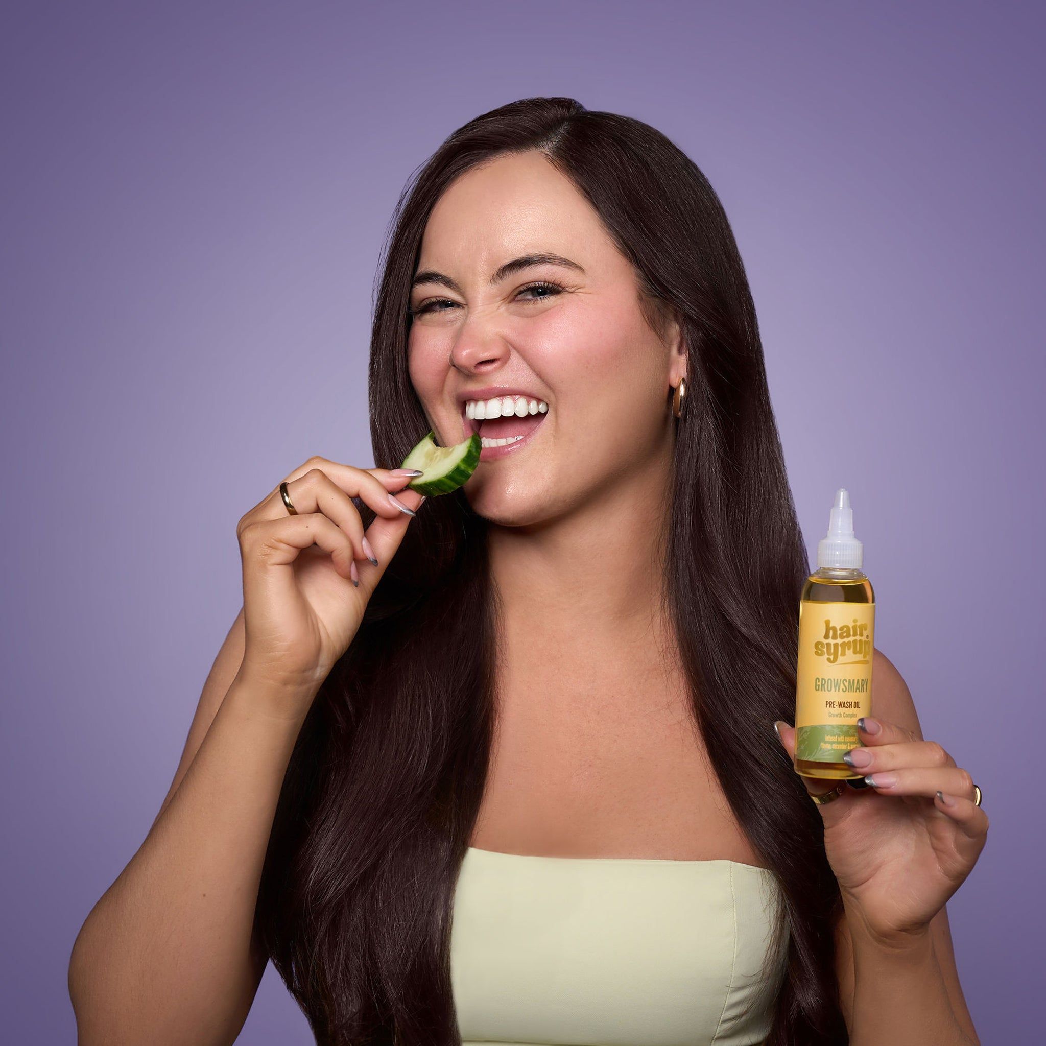 Frau mit langen, dunklen Haaren, isst Gurke. Hält eine gelbe Flasche mit Etikett.