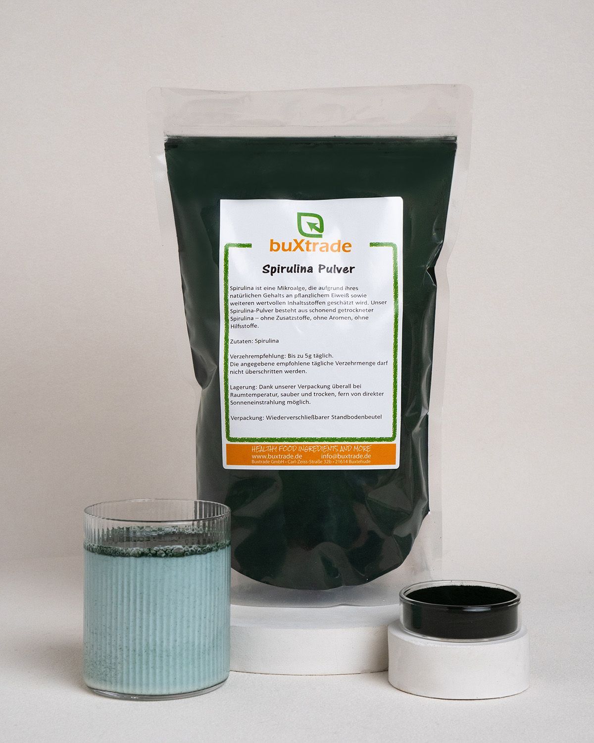 Verpackung von buXtrade Spirulina-Pulver, ein Glas Getränk und ein Behälter. Das Getränk ist blau.