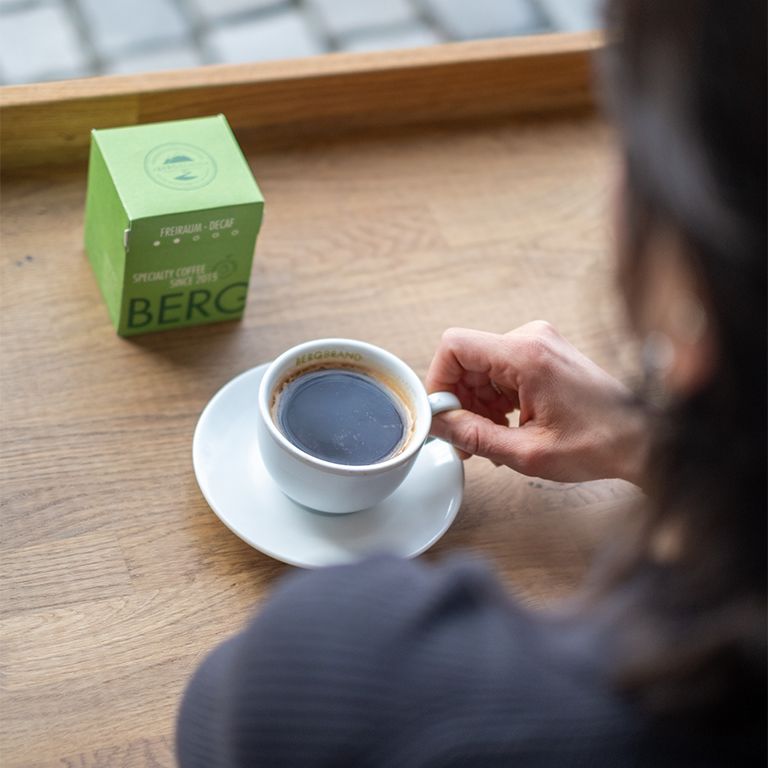 Kaffeetasse mit Kaffee und Verpackung im Hintergrund. BERGBRAND-Logo auf der Tasse. Holzoberfläche.