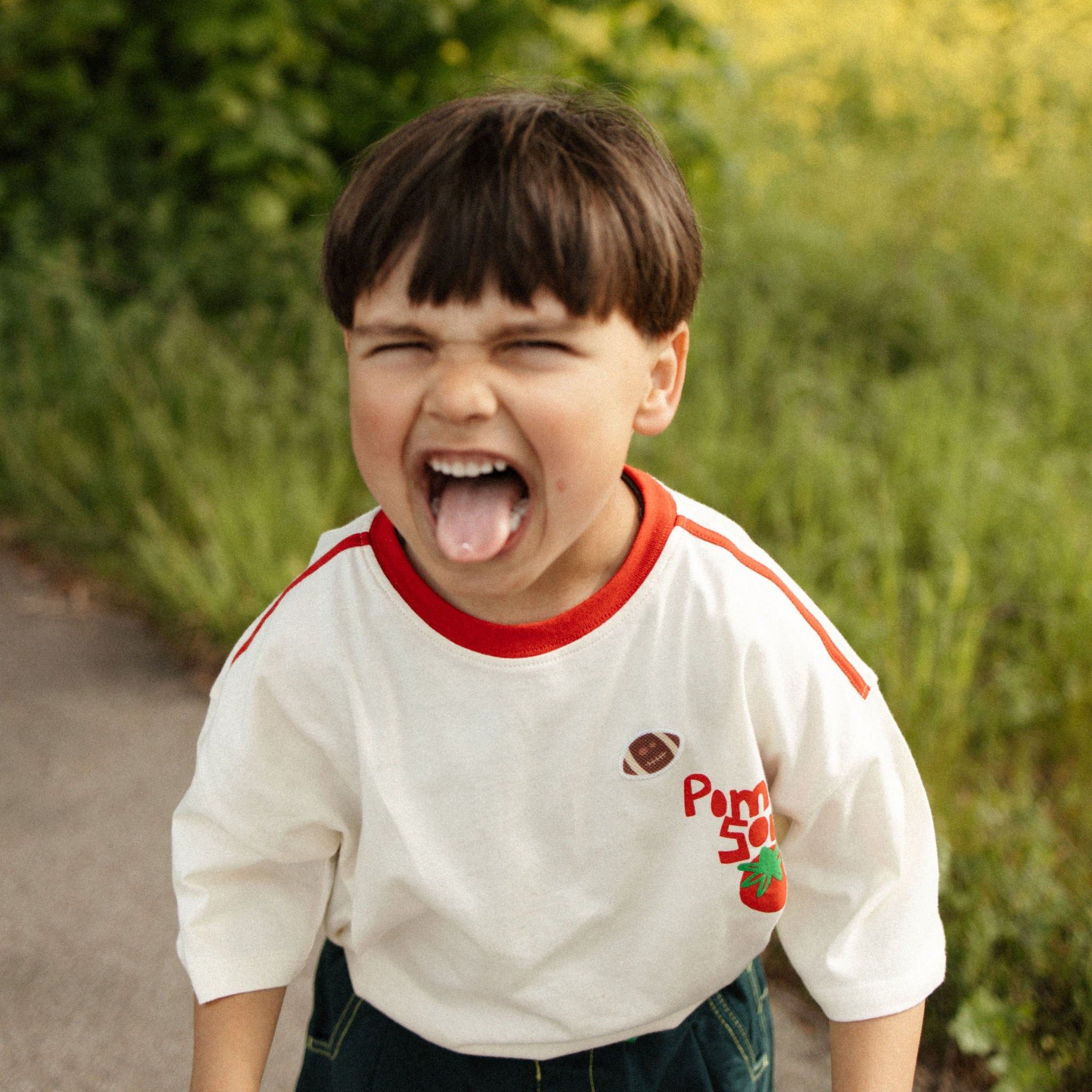 Junge mit gestreiftem Shirt, das mit Erdbeere und Football verziert ist, streckt Zunge heraus.