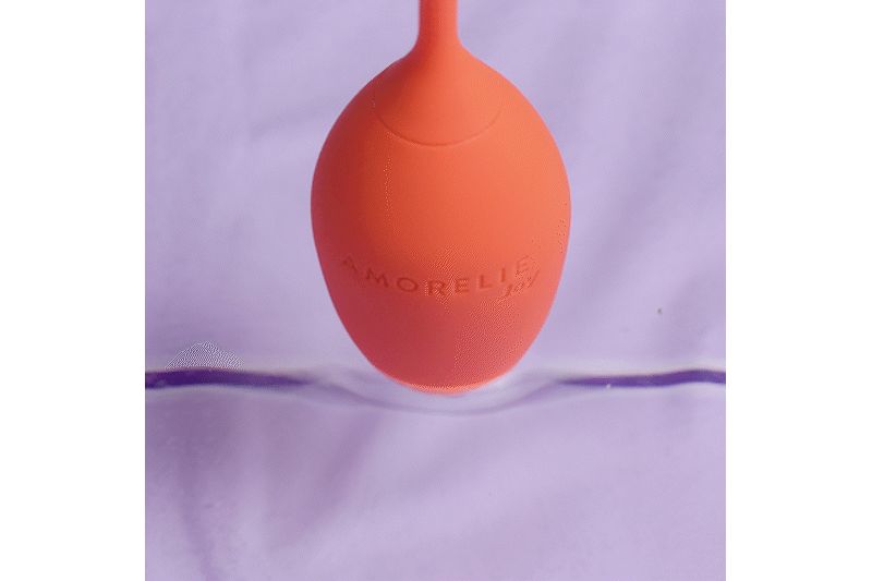 Orangener, eiförmiger Gegenstand mit der Aufschrift AMORELIE Joy. Der Gegenstand befindet sich in einer Flüssigkeit.