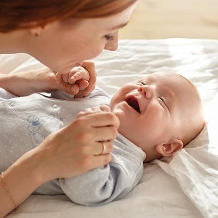 Mutter und Baby. Baby liegt auf dem Rücken, lächelt. Mutter beugt sich vor, hält Hände des Babys.