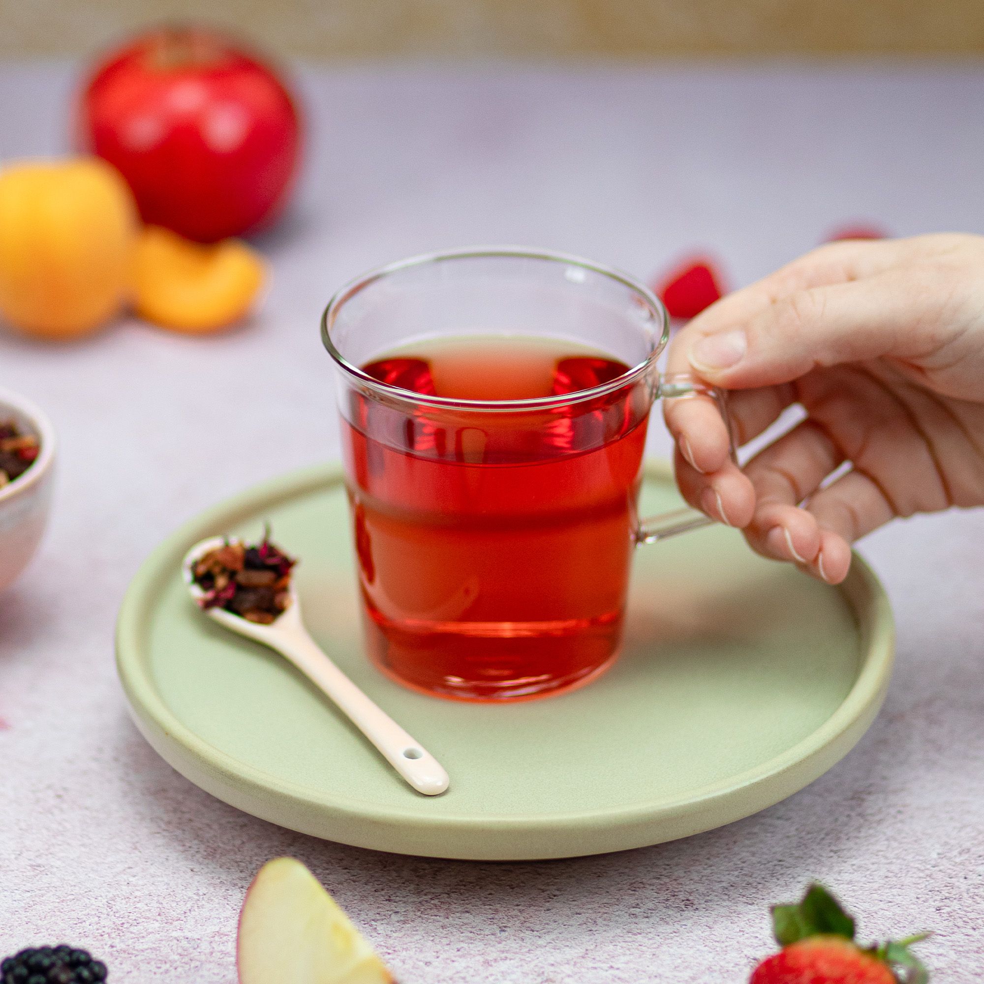 Glas mit rotem Getränk, auf Untersetzer. Löffel mit Teemischung daneben. Hand hält Glas. Früchte im Hintergrund.