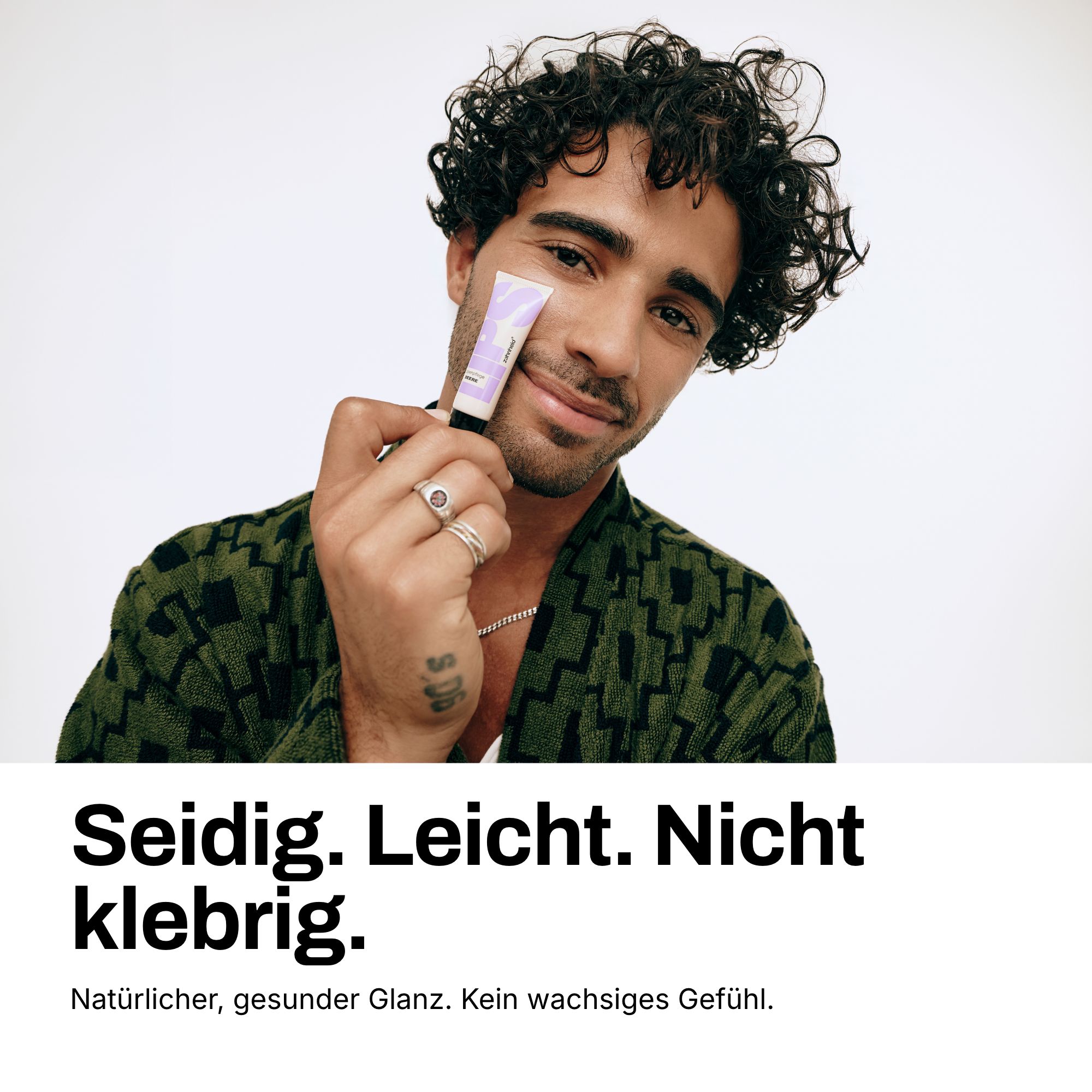 Mann mit lockigem Haar hält eine Lippenpflege-Tube. Text: Seidig. Leicht. Nicht klebrig.