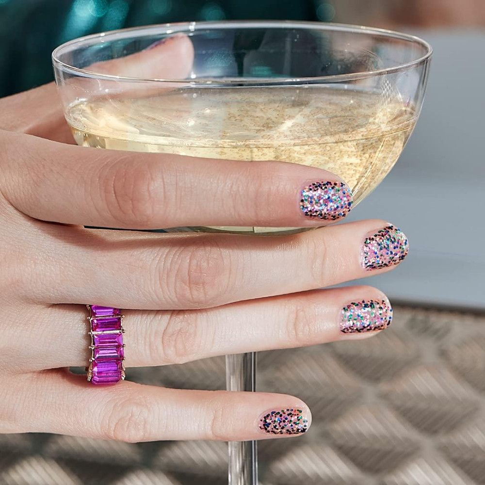 Hand mit lackierten Nägeln, die ein Glas Champagner hält. Nagellack mit bunten Glitzerpartikeln.