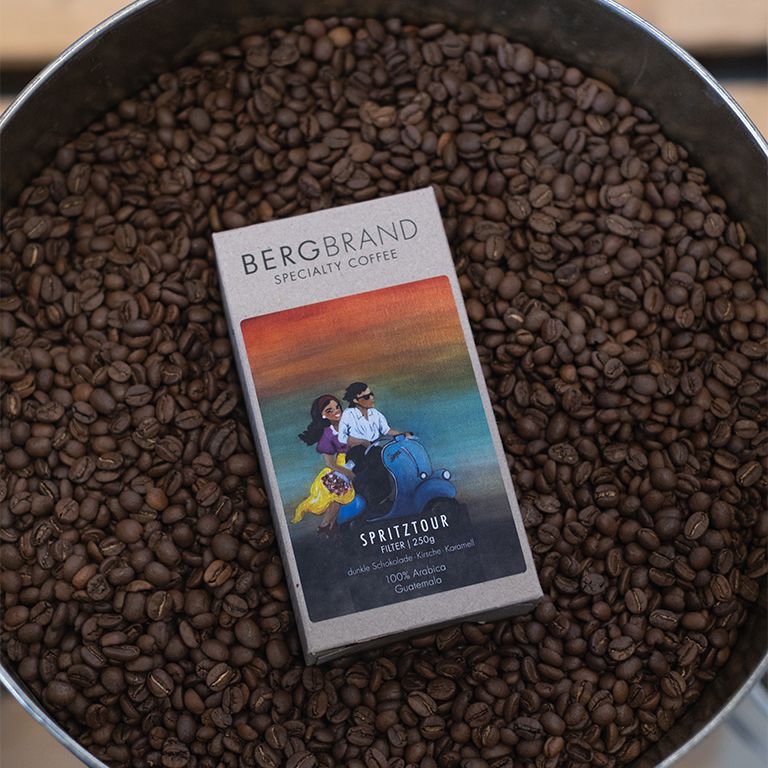 Kaffeebohne-Hintergrund mit einer Packung BERGBRAND Specialty Coffee Spritztour. Karton mit Roller-Illustration.