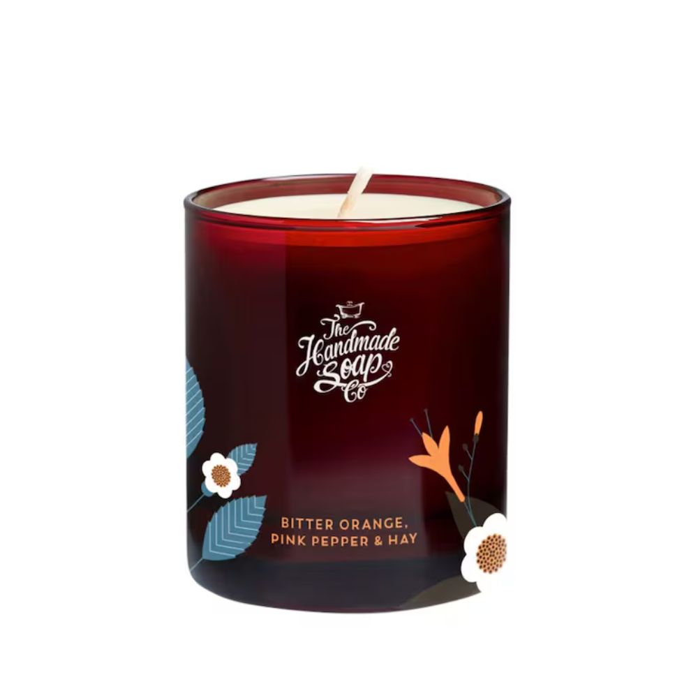 Runde Kerze im Glas. Dunkelrotes Glas mit floralen Mustern und Text: Bitter Orange, Pink Pepper & Hay. Marke: The Handmade Soap Co.