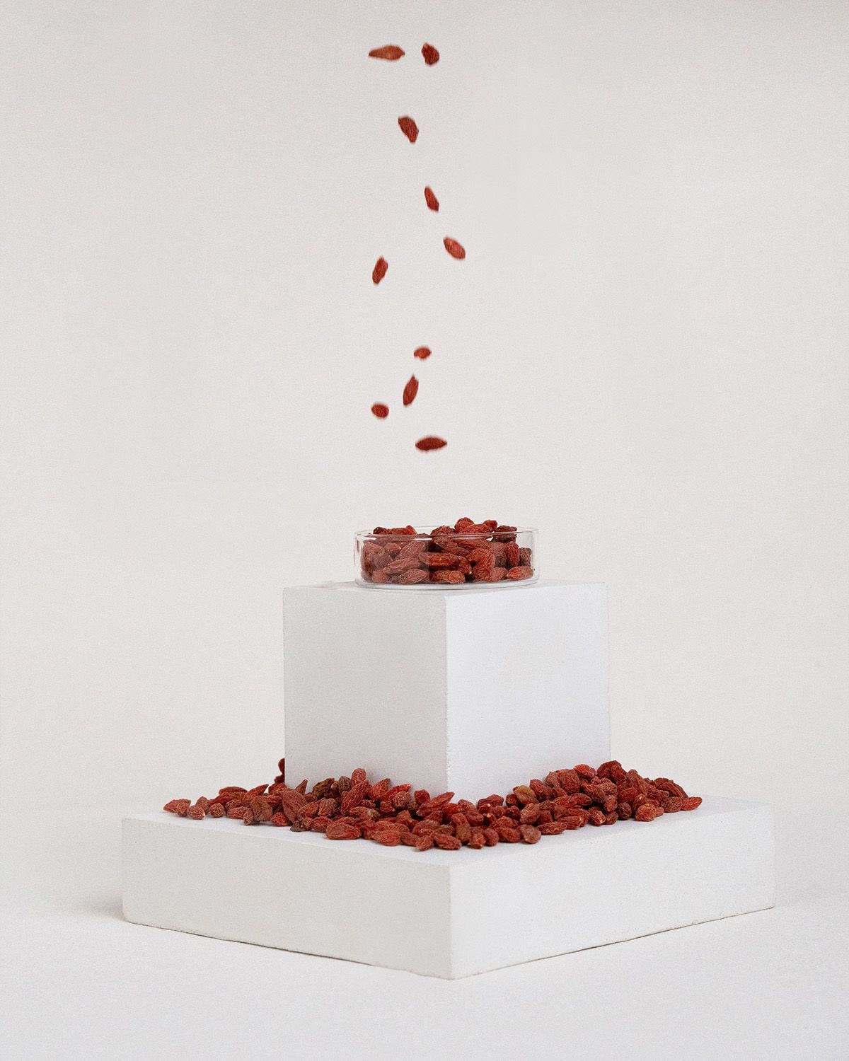 Gojibeeren fallen in eine Schale auf einem weißen Podest. Mehr Beeren liegen darunter.
