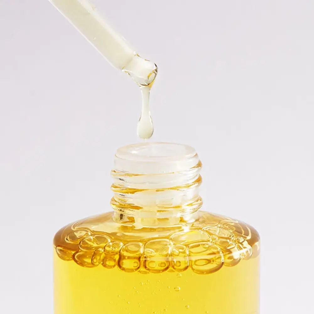 Ein Tropfen goldgelbes Serum fällt aus einer Pipette in eine Glasflasche mit ähnlicher Flüssigkeit und Luftblasen.