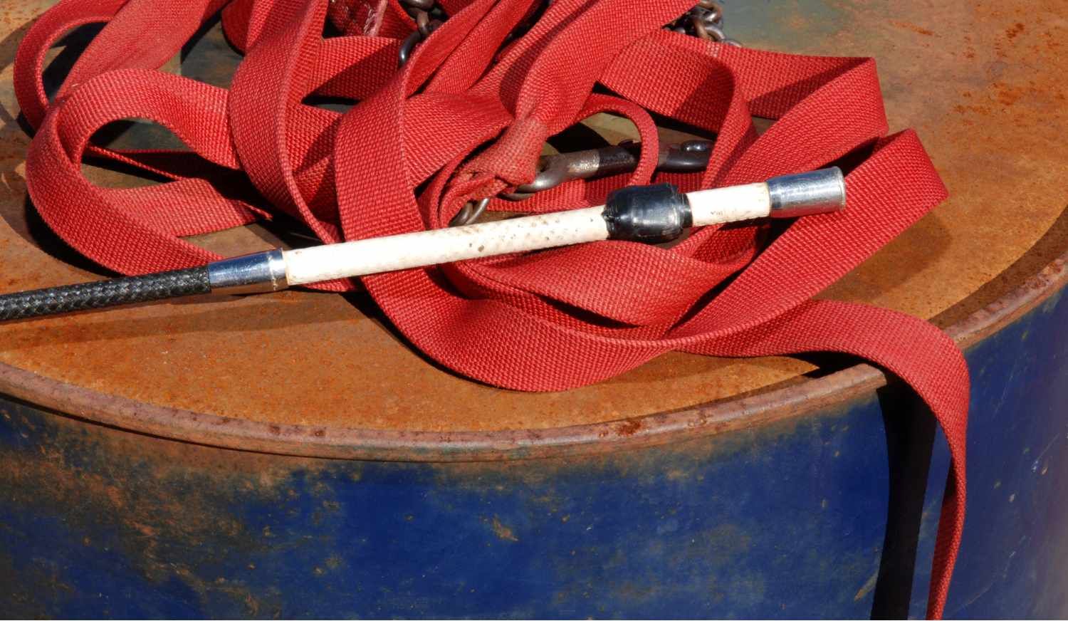 Reitgerte mit rotem Gurtband auf einem rostigen Fass. Gerte mit weißem Mittelteil und Metallverbindungen.