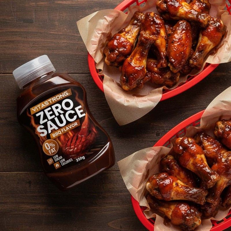 Braune Flasche mit weißem Deckel. Aufschrift: Vitastrong Zero Sauce BBQ Flavor. 0% Fett, Low Sugar, Low Calories. 250g. Neben zwei Körben mit Hähnchenflügeln.