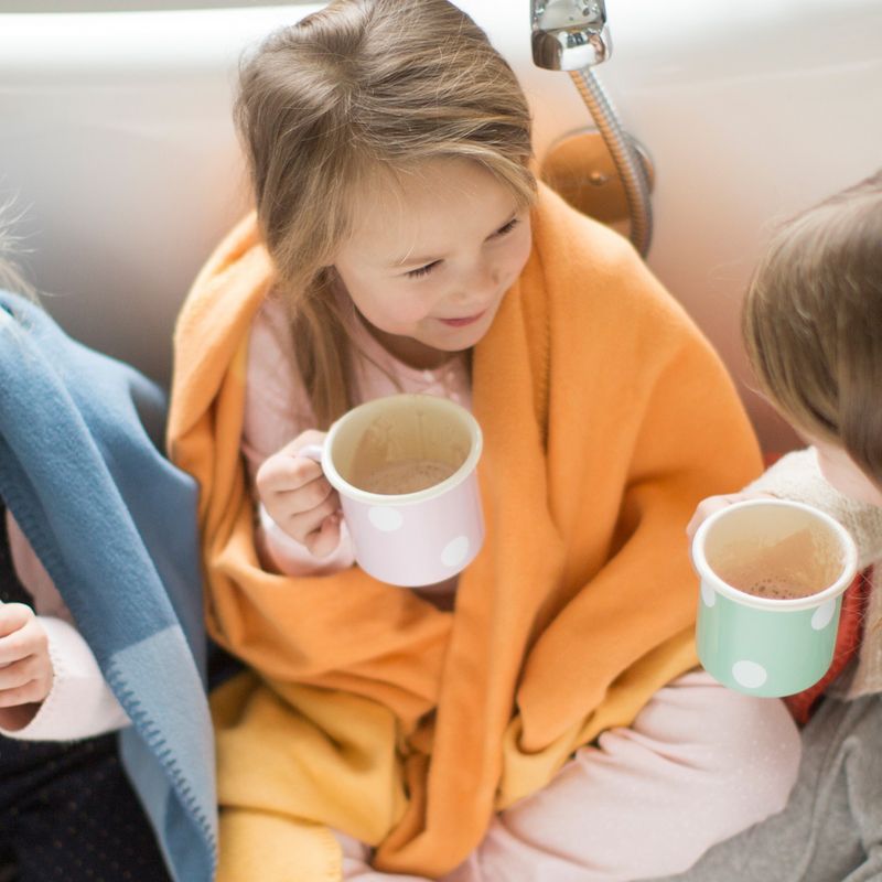 Kind mit orangefarbener Decke und Tasse. Zwei weitere Kinder mit Decken und Tassen. Badezimmerumgebung.