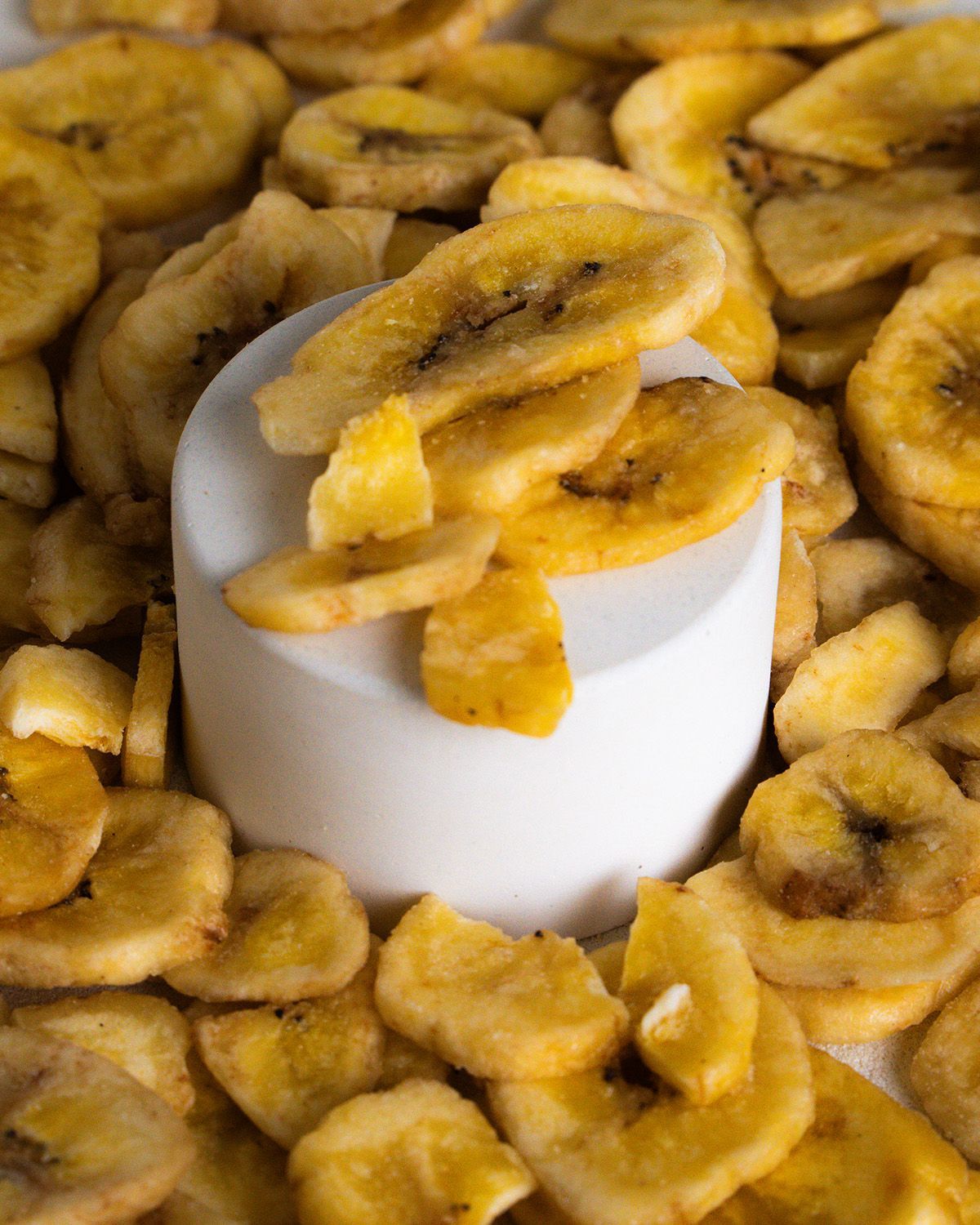 Nahaufnahme von Bio Bananenchips auf einem weißen Podest, umgeben von Chips. Gelbliche, runde Chips mit dunklen Punkten.