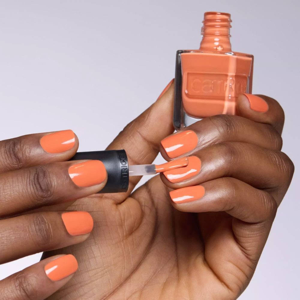 Hand mit lackierten Nägeln in Orange. Nagellackflasche mit Pinsel. Catrice-Logo sichtbar.