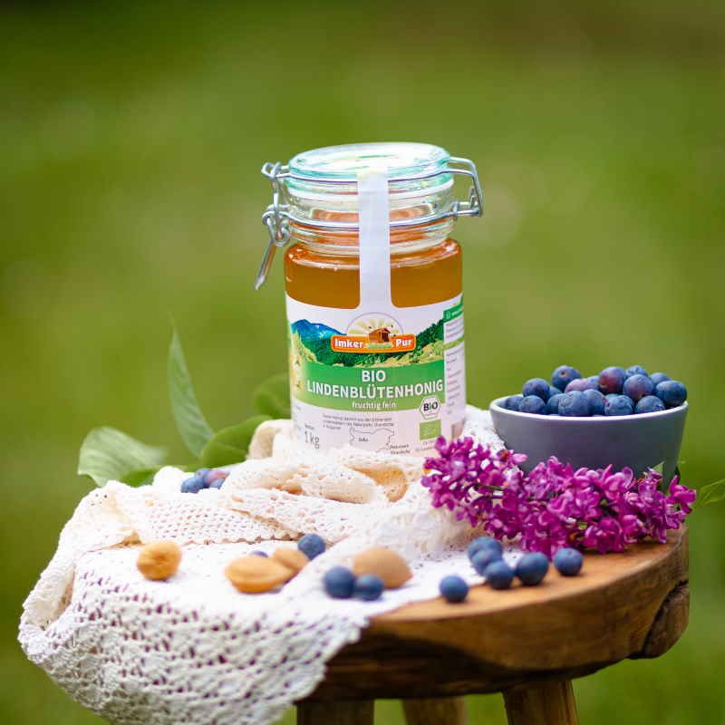 Honigglas "ImkerPur Bio Lindenblütenhonig" auf Holzschemel. Blaubeeren, Blüten und Kekse daneben.
