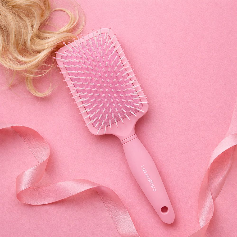 Rosa Paddle Brush mit weißen Borsten. Der Griff trägt den Markennamen. Blondes Haar und rosa Band im Hintergrund.