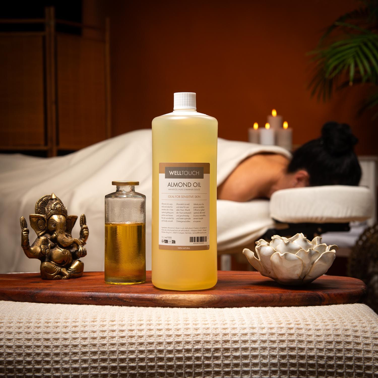 Große Flasche Mandelöl, daneben kleinere Flasche. Auf Holztablett, im Hintergrund Person auf Massageliege.