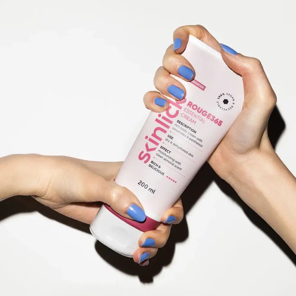 Hände halten Creme-Tube SKINLICK Rouge365 Essential Cream. Rosa-weißes Design, roter Streifen. Blaue Fingernägel. Text sichtbar.
