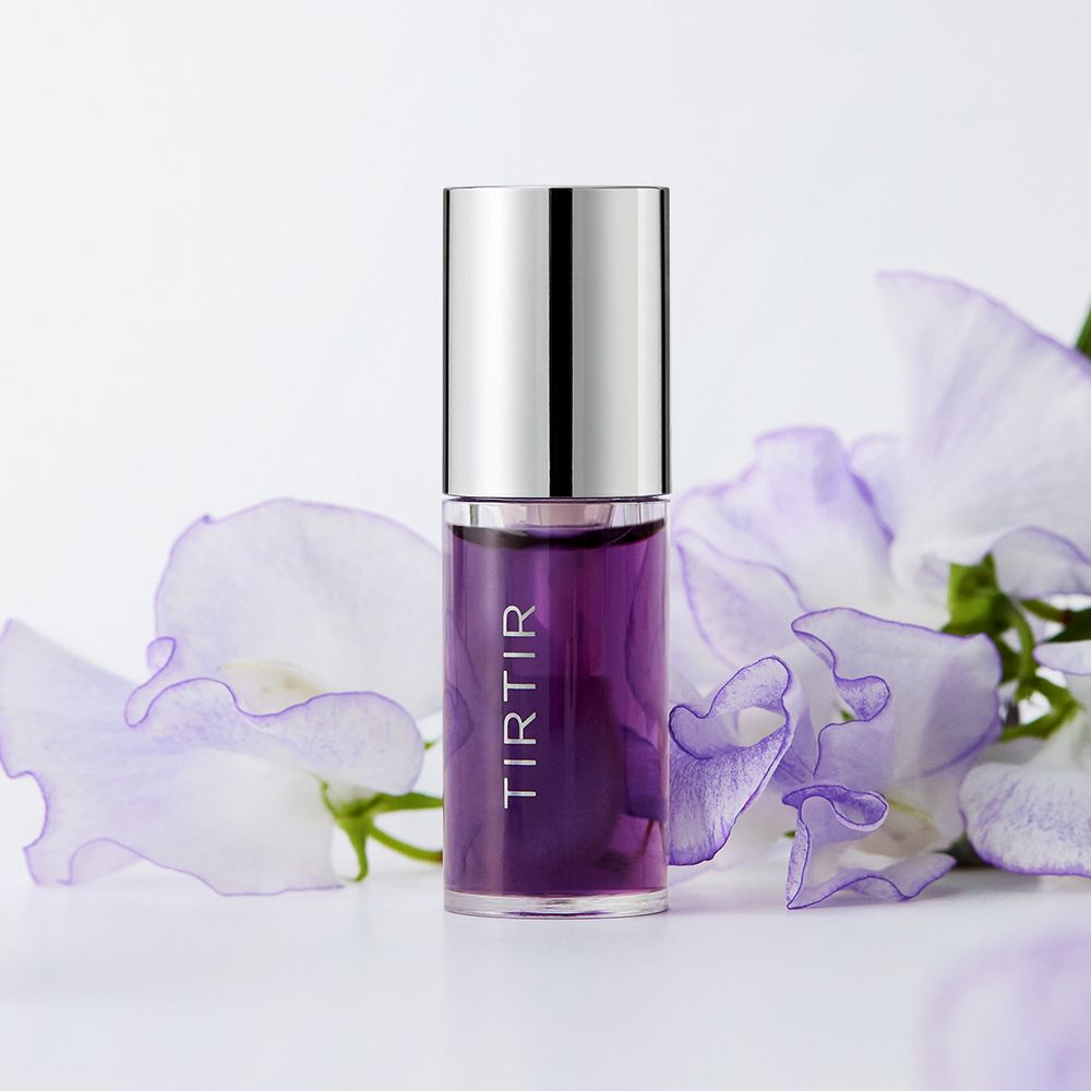 TIRTIR Lip Oil Lavender. Violettes Öl in einer transparenten Flasche mit silbernem Deckel. Blütenblätter im Hintergrund.