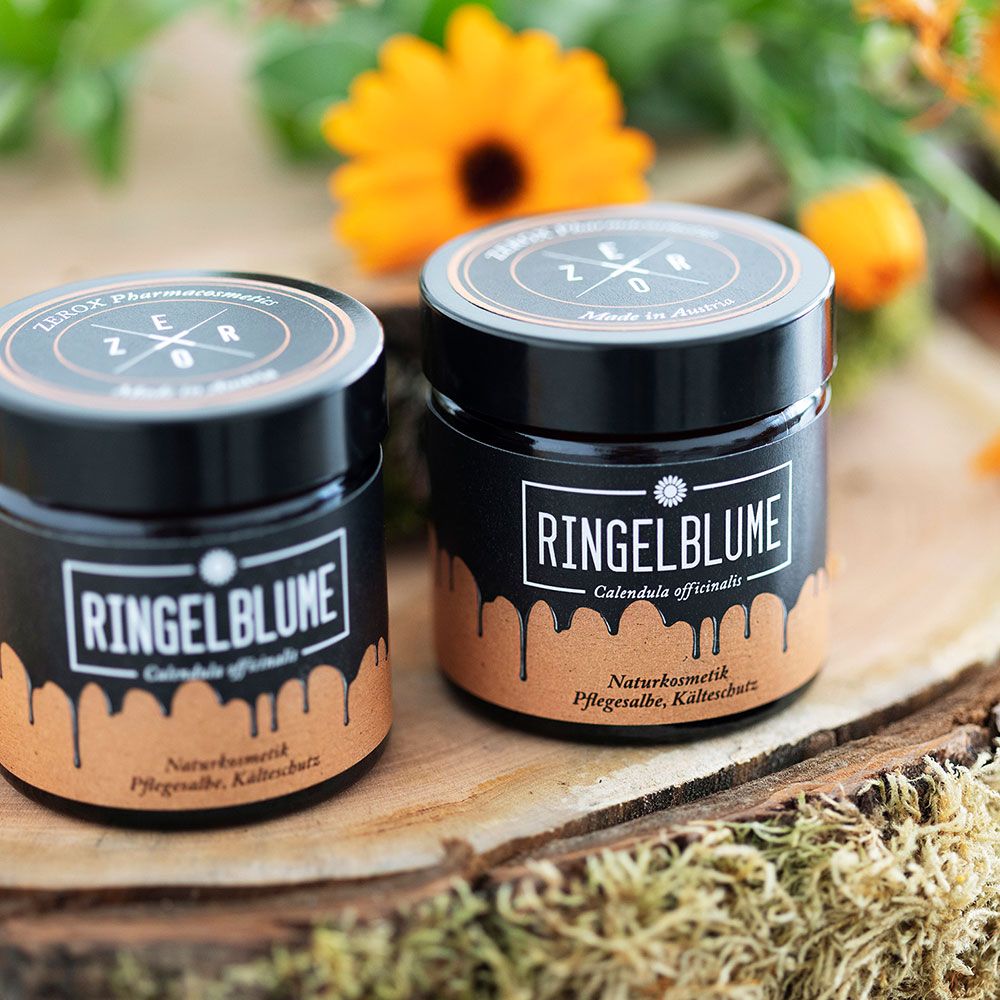 Zwei Gläser mit schwarzem Deckel. Etikett "Ringelblume" und "Calendula officinalis". Auf Holzbrett mit Blüte im Hintergrund.