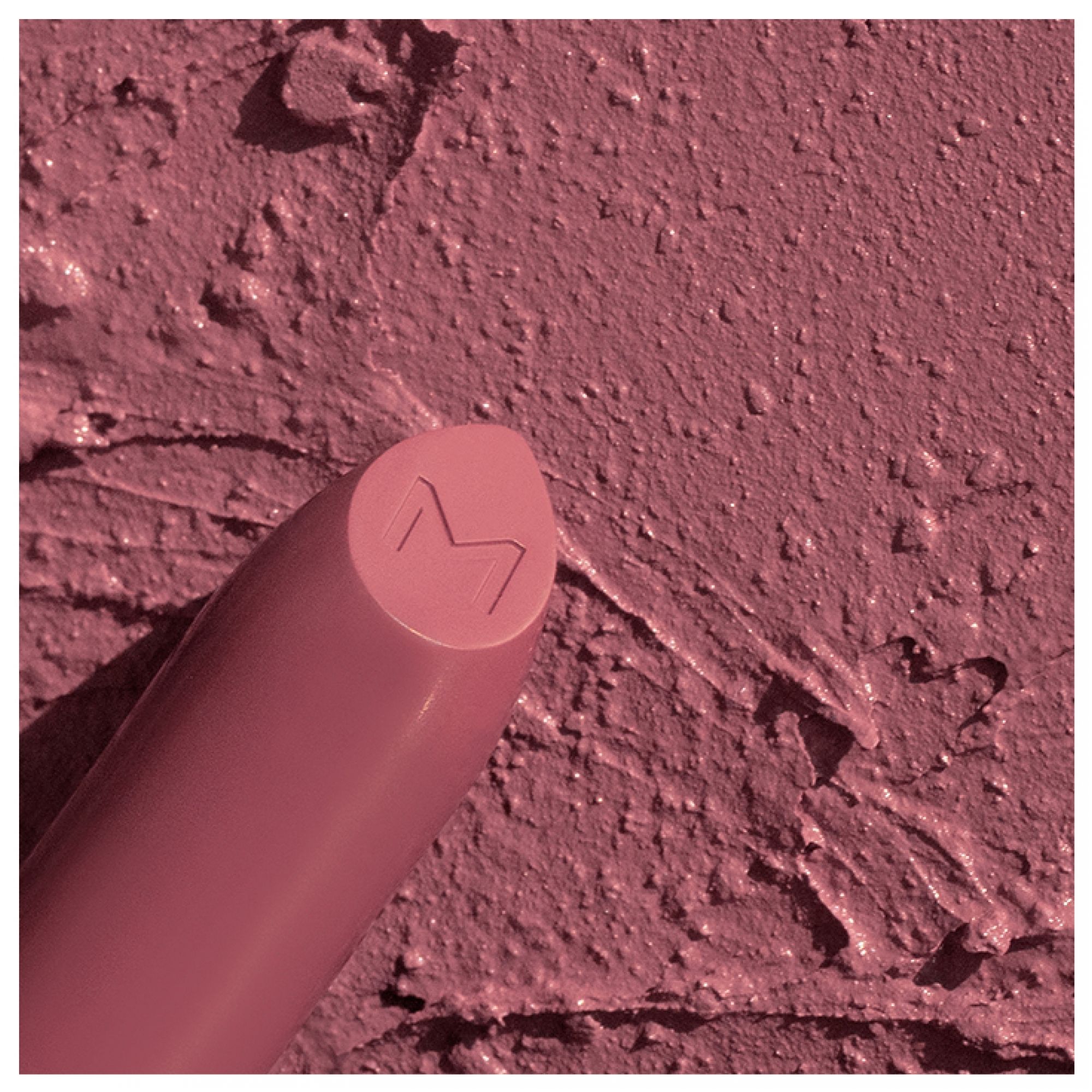 Ein matter, rosafarbener Lippenstift mit eingraviertem 'M'-Logo auf der Spitze, auf einer Textur aus demselben Produkt.
