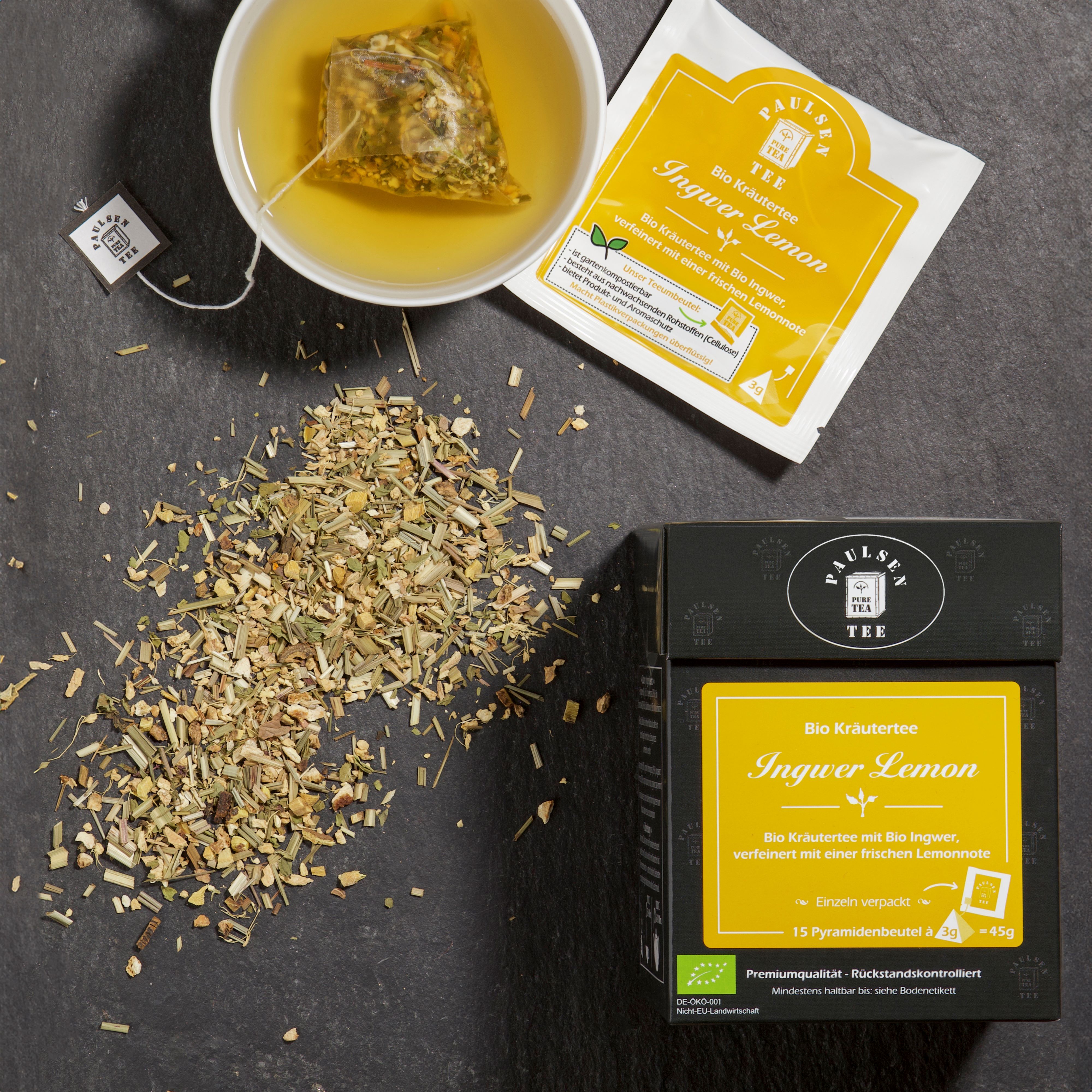 Tasse Tee mit Teebeutel. Daneben Teebeutel-Päckchen und Box mit Aufschrift: Bio Ingwer Lemon Tee. Lose Teeblätter.