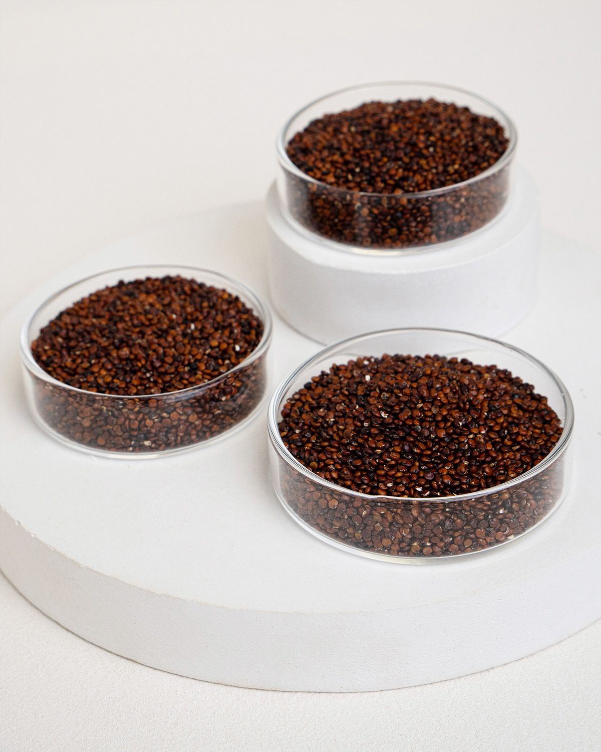 Drei runde, transparente Schalen mit rotem Quinoa auf weißem Podest. Eine Schale oben, zwei darunter.