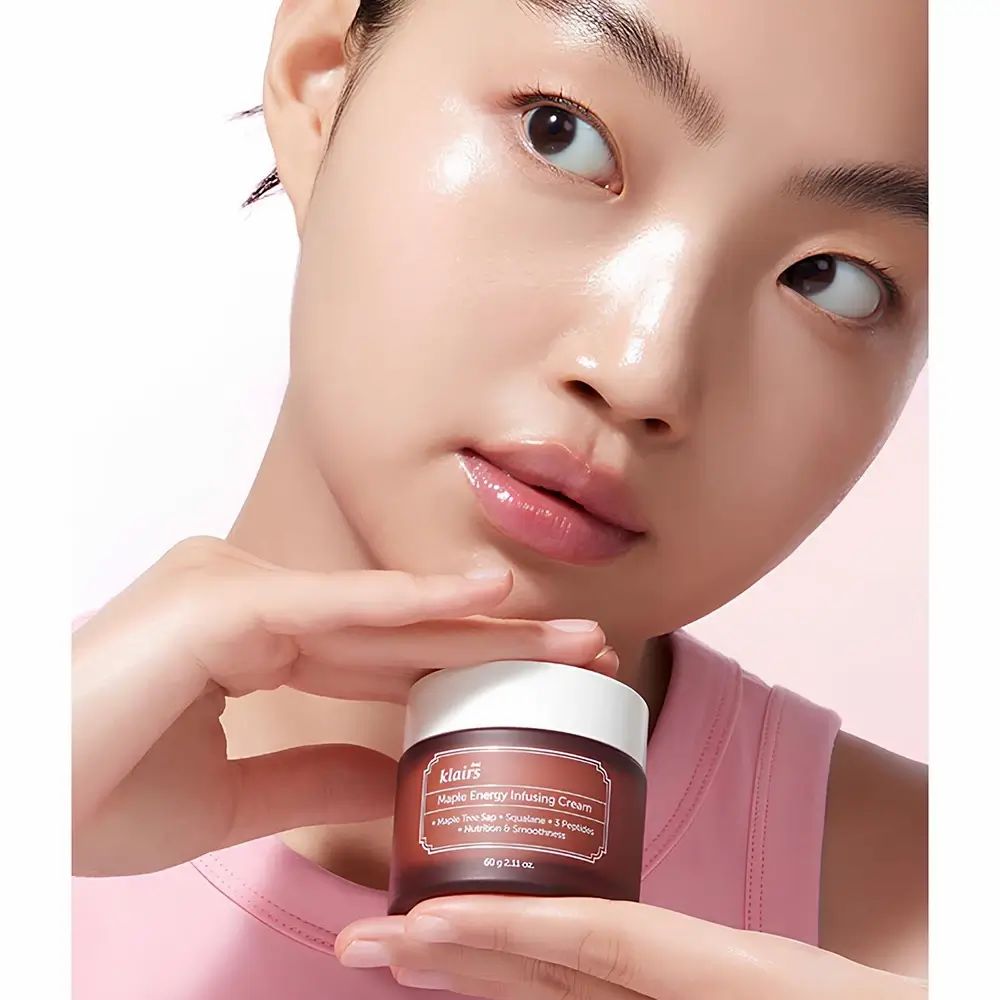 Frau hält eine braune Creme-Dose. Aufschrift: Dear Klairs Maple Energy Infusing Cream. Gesicht glänzt. Rosa Oberteil.