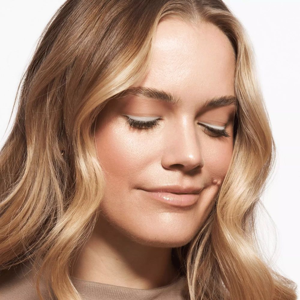 Porträt einer Frau mit blonden Locken, die die Augen geschlossen hat. Sie trägt hellen Eyeliner und Lippenbalsam.