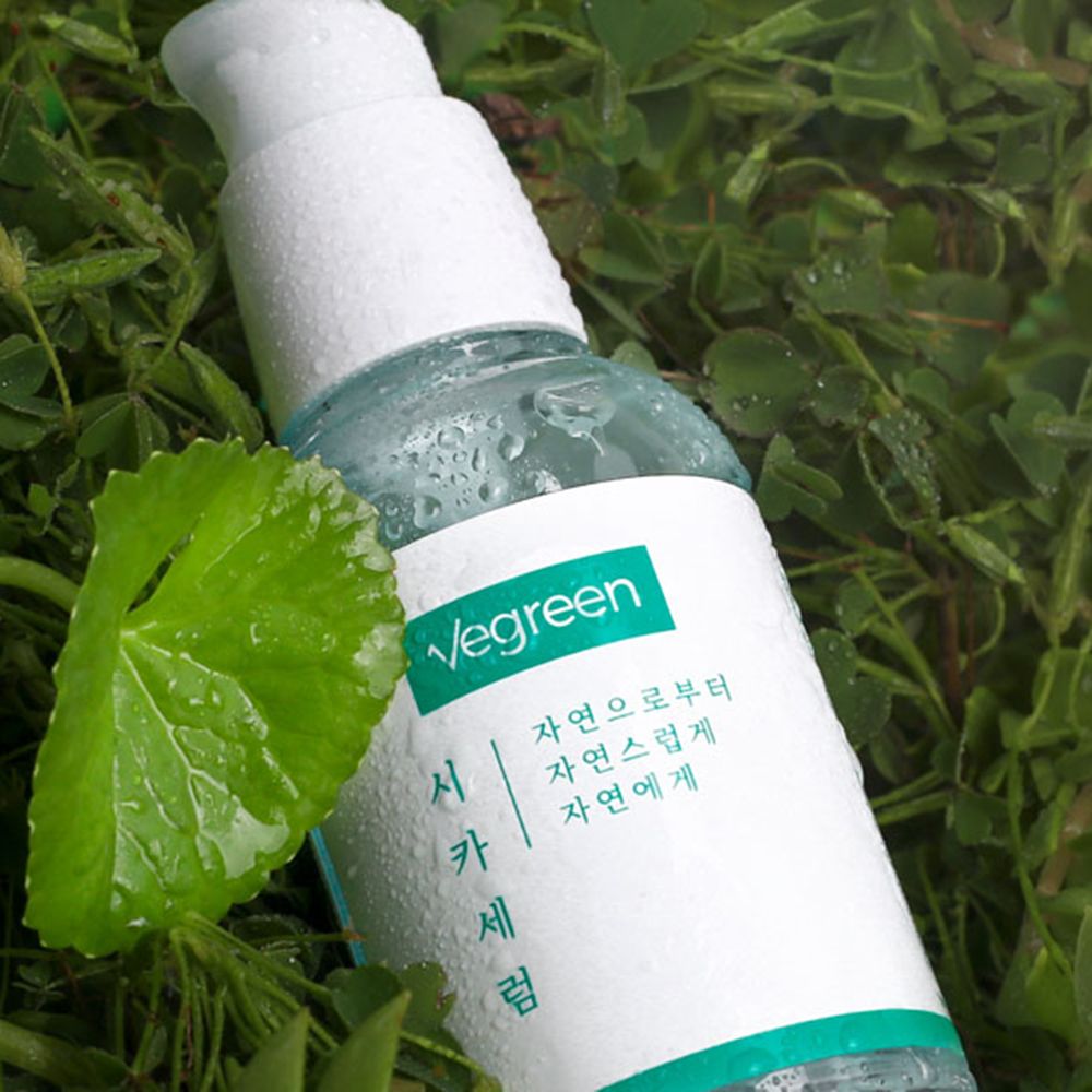 Gesichtsserum-Flasche im Gras. Weißer Pumpdeckel, grünes Etikett mit Text. Tropfen auf der Flasche und einem Blatt.