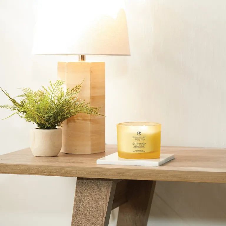 Gelbe Kerze auf einem Tisch. Neben einer Lampe und einer Topfpflanze. Aufschrift: 'Chesapeake Bay Mind & Body'.