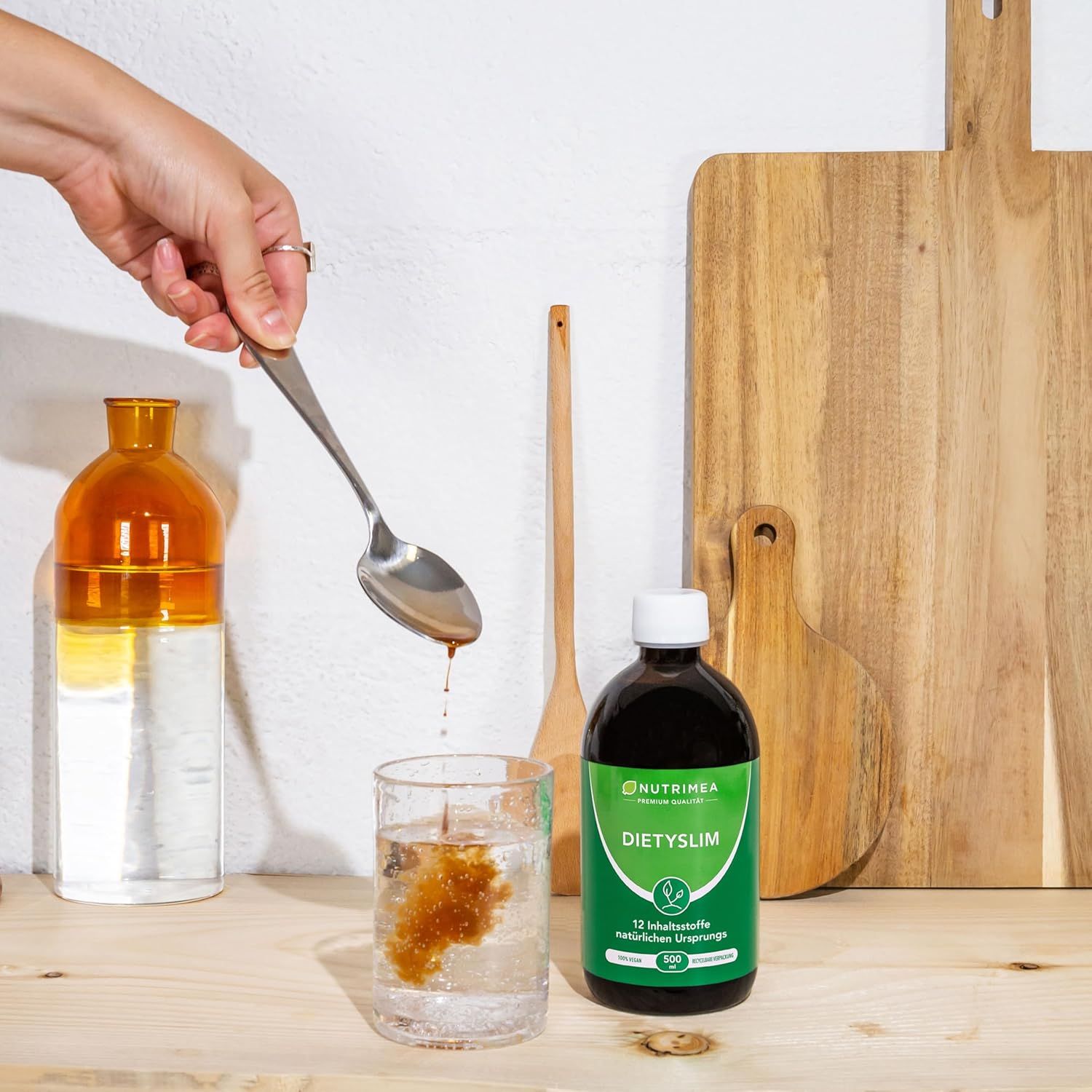 Hand mit Löffel über Glas. Flasche mit grünem Etikett: DIETYSILM. Auf Holzbrett und in Glas. Wasser und Flasche.