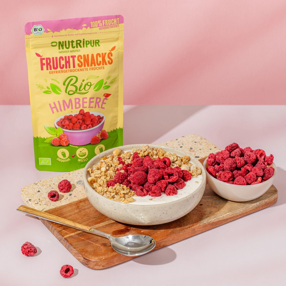 Beutel mit gefriergetrockneten Bio-Himbeeren, Schale mit Himbeeren, Schale mit Müsli und Himbeeren, Holzteller mit Löffel.