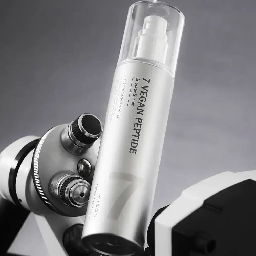 Silberne Flasche mit Sprühkopf und klarer Kappe. Aufschrift "7 Vegan Peptide Booster Serum". Flasche liegt auf einem Mikroskop.