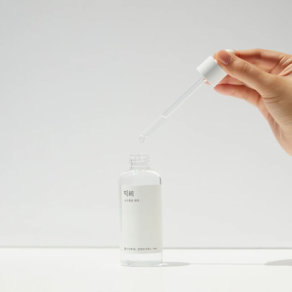 Hand hält Pipette über offener Flasche, ein Tropfen fällt heraus. Klare Essenz.