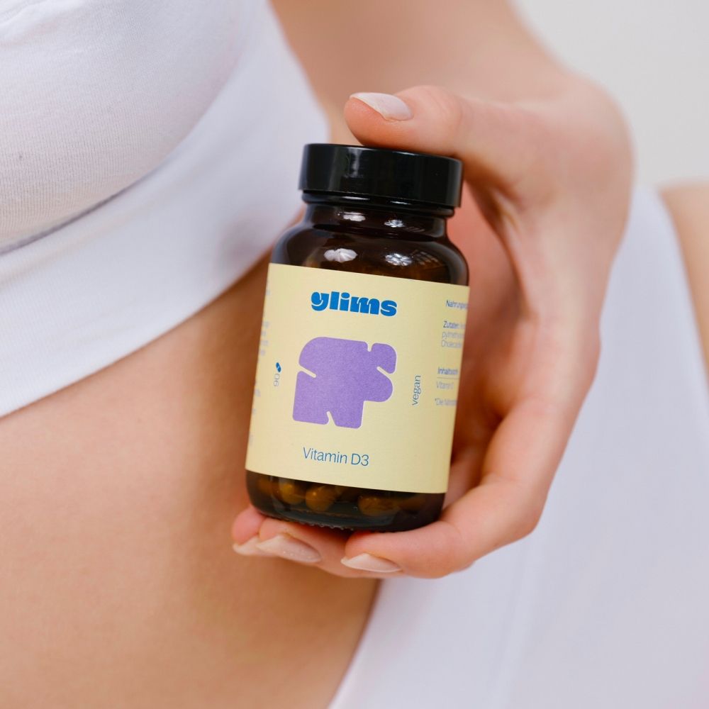 Person hält eine braune Glasflasche mit schwarzem Deckel. Etikett mit "glims"-Logo, lila Form und "Vitamin D3".