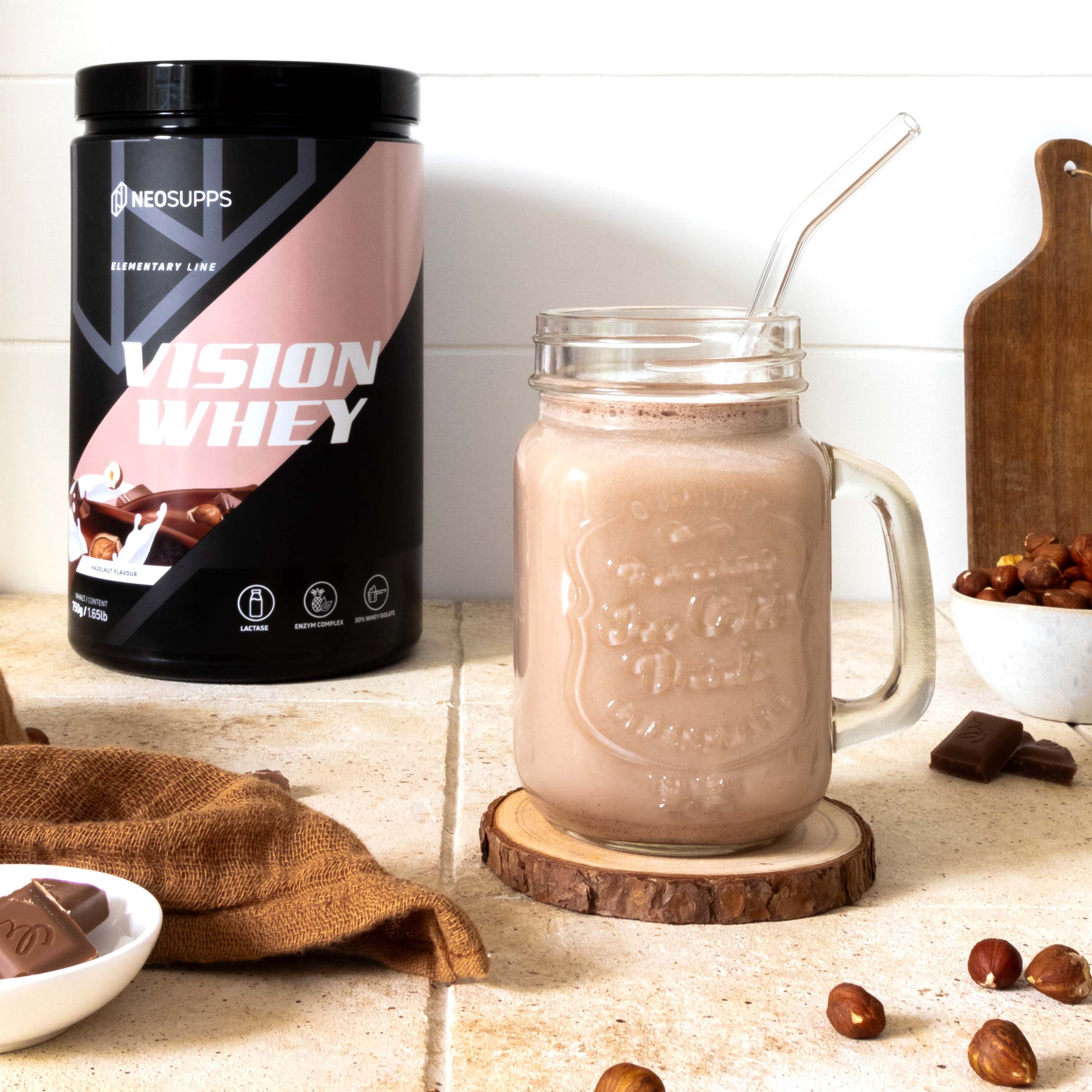 Glas mit Shake, Dose Vision Whey. Dose schwarz, rosa Akzent. Text: Vision Whey, Neosupps. Haselnuss-Stückchen, Schokolade, Holzuntersetzer.