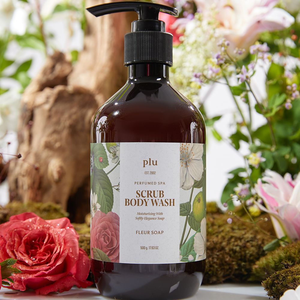 Braune Flasche mit Pumpe, floralem Design und Text: SCRUB BODY WASH, FLEUR SOAP. Marke: plu. Auf Blumen und Holz platziert.