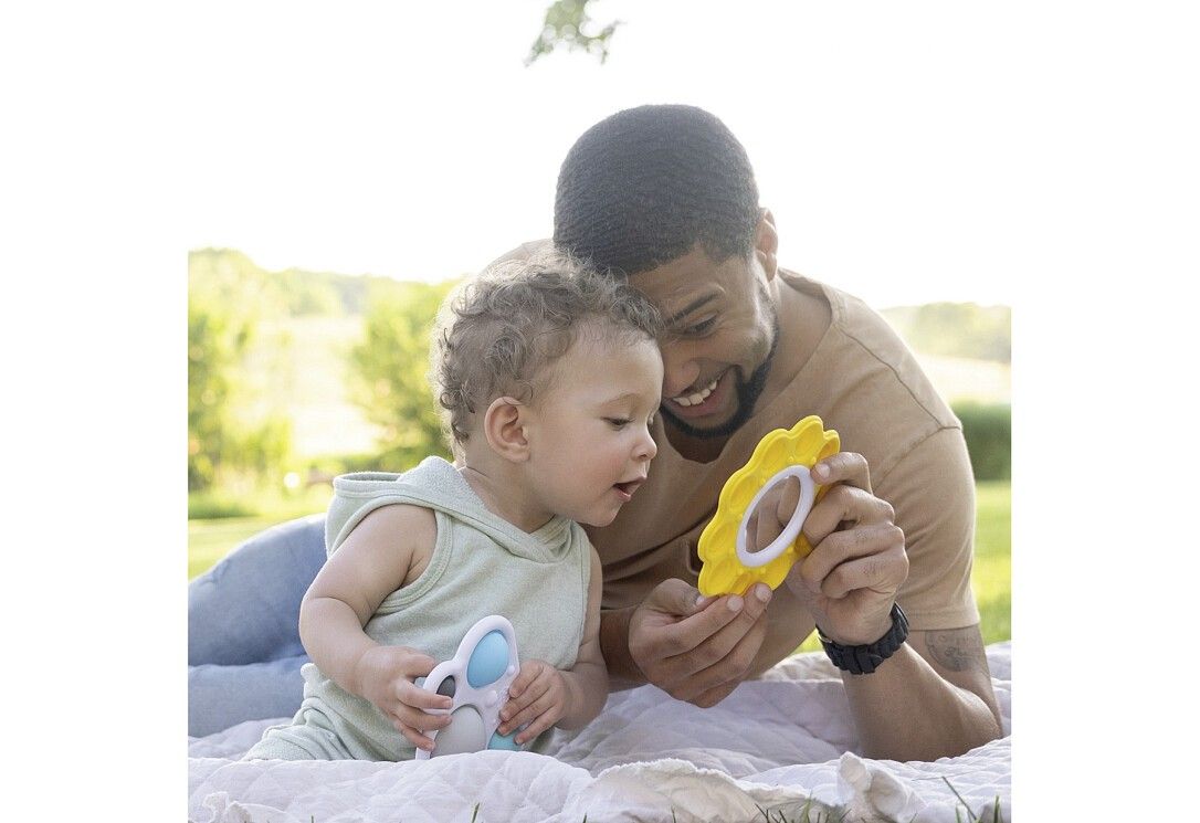 Ein Mann und ein Kind sitzen im Freien. Das Kind hält ein blau-weißes Spielzeug, der Mann ein gelbes, blütenförmiges Spielzeug.